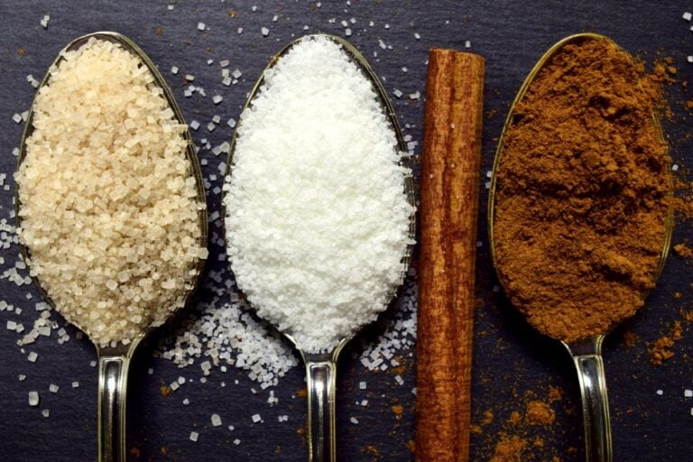 Three spoons with brown sugar, white sugar, and cinnamon powder sit alongside a cinnamon stick on a dark surface, showcasing an array of flavored sugars.
