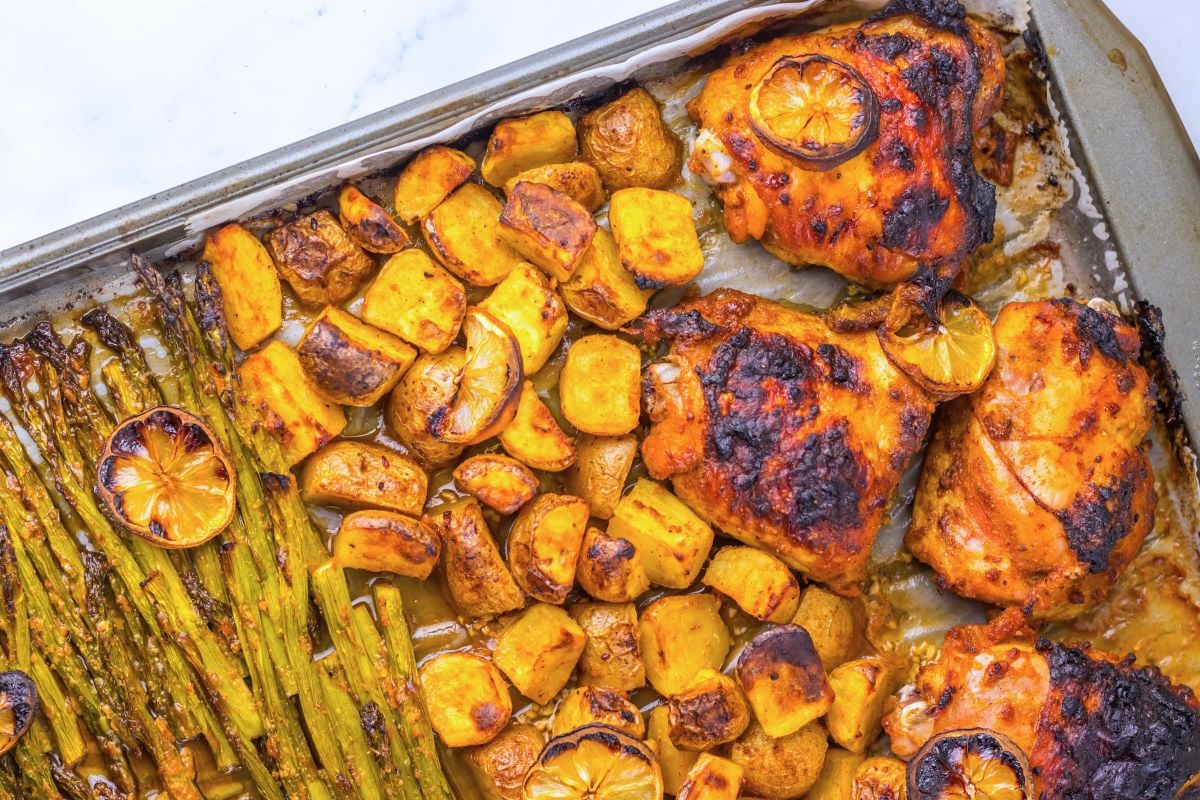Sheet pan with roasted chicken thighs, lemon slices, asparagus, and diced potatoes, all cooked to a golden brown.