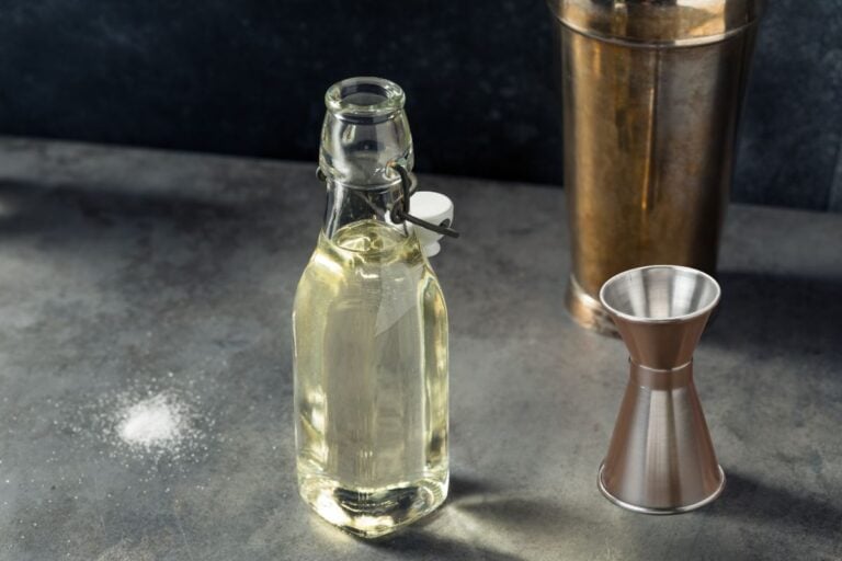 A glass bottle filled with clear liquid sits next to a metal jigger and a cocktail shaker on a gray countertop, with some spilled salt nearby.