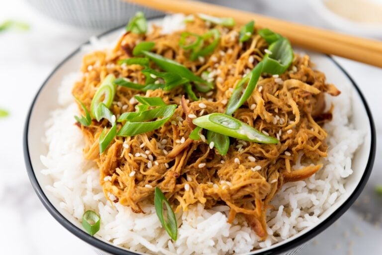 A bowl of white rice topped with shredded chicken, green onions, and sesame seeds, with chopsticks in the background.