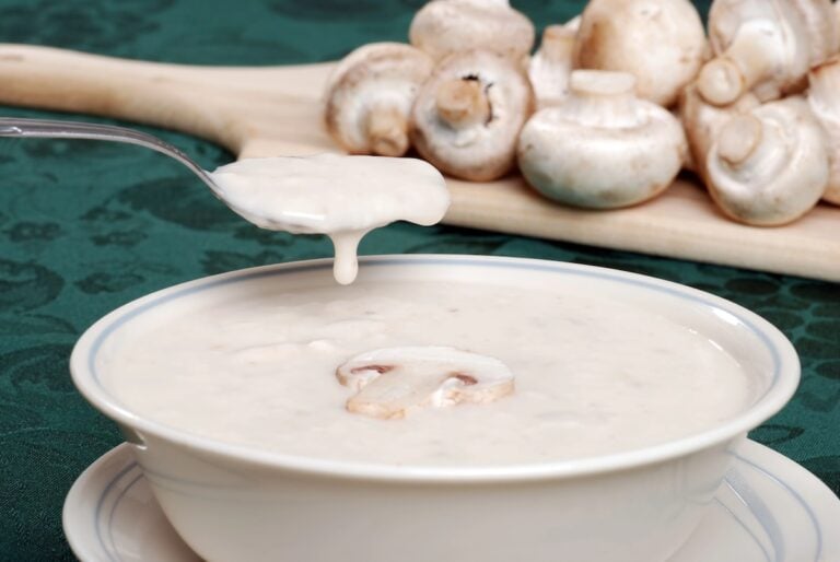 A bowl of creamy mushroom soup with sliced mushrooms, and a spoon drizzling soup back into the bowl. A cutting board with whole mushrooms is in the background.