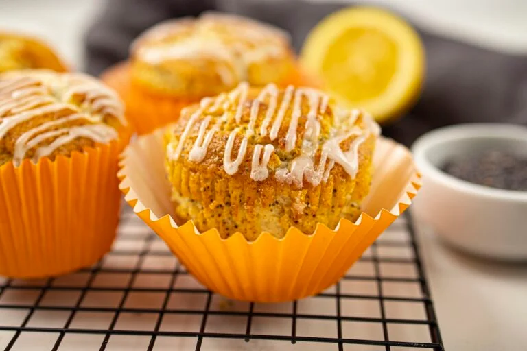 A lemon poppy seed muffin with white icing sits in an orange wrapper on a cooling rack. A lemon slice and poppy seeds in a small dish are in the background.