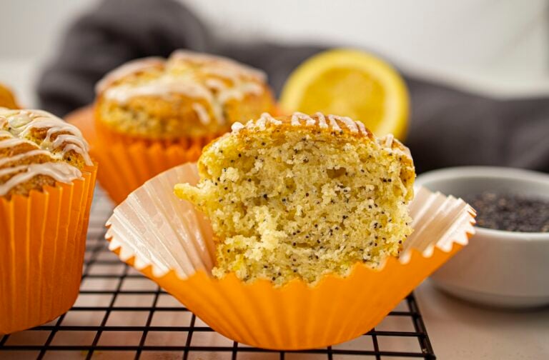 Lemon poppy seed muffins, one partially eaten, sit on a cooling rack with orange wrappers. A lemon wedge and a bowl of poppy seeds are in the background.