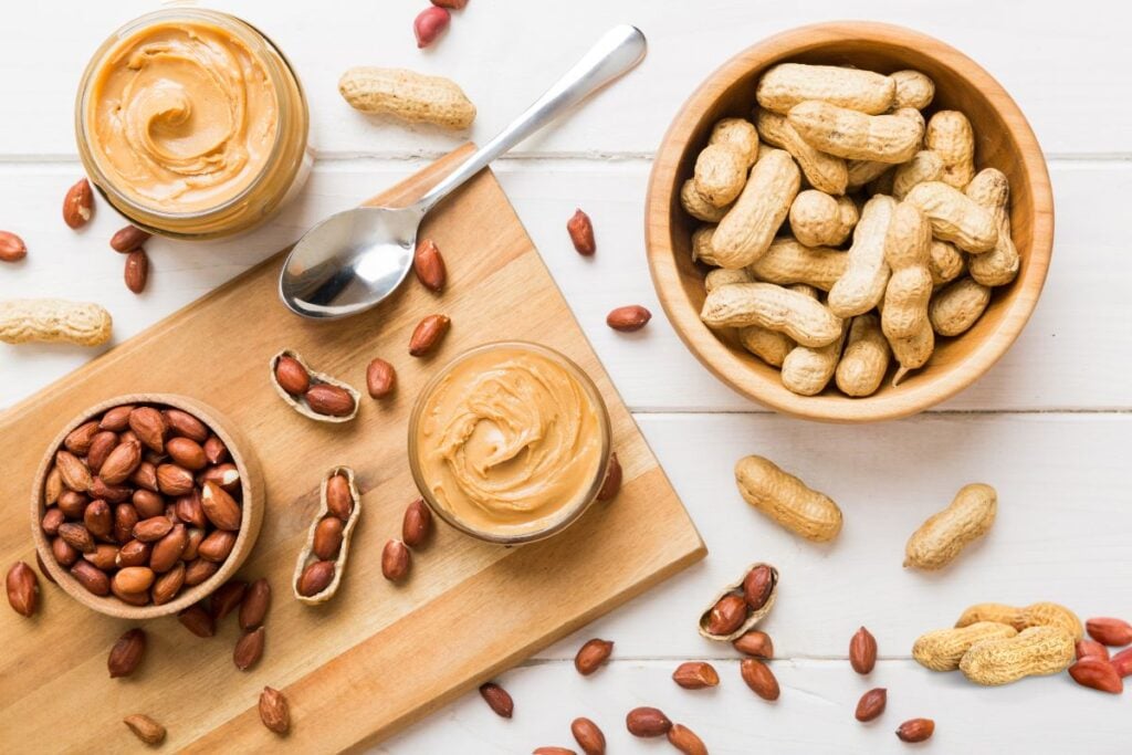 Bowls of whole peanuts, shelled peanuts, and peanut butter jars on a wooden board, with a metal spoon and scattered peanuts on a white surface.