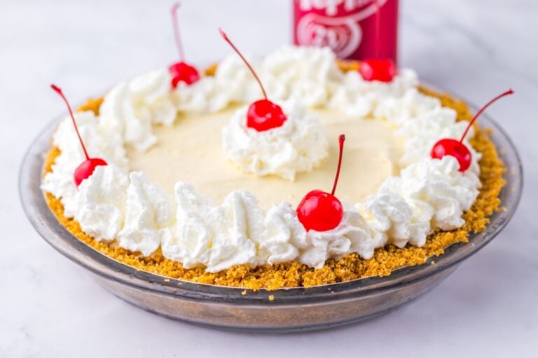 A Dr. Pepper pie with a graham cracker crust, topped with whipped cream and cherries, is displayed on a white surface.