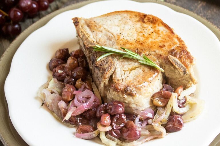 A cooked pork chop, perfect for Easter recipes, served on a plate with caramelized onions, grapes, and garnished with a sprig of rosemary.