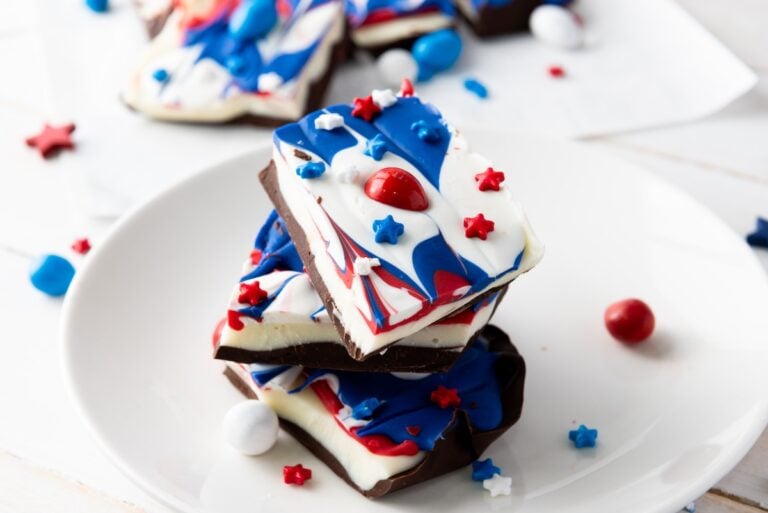 A stack of patriotic-themed chocolate bark with red, white, and blue swirls, topped with star-shaped sprinkles and candy, on a white plate.