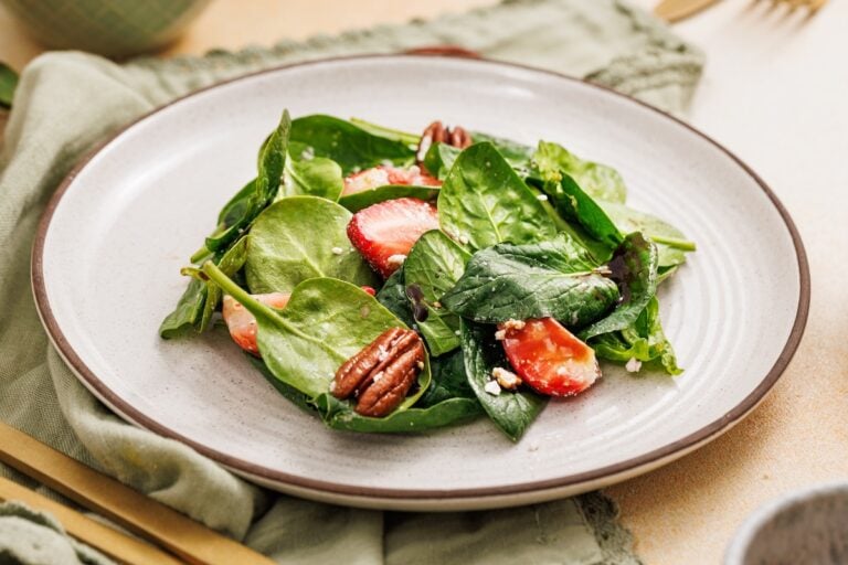 A salad with fresh spinach leaves, sliced strawberries, pecans, and crumbled cheese on a white plate.