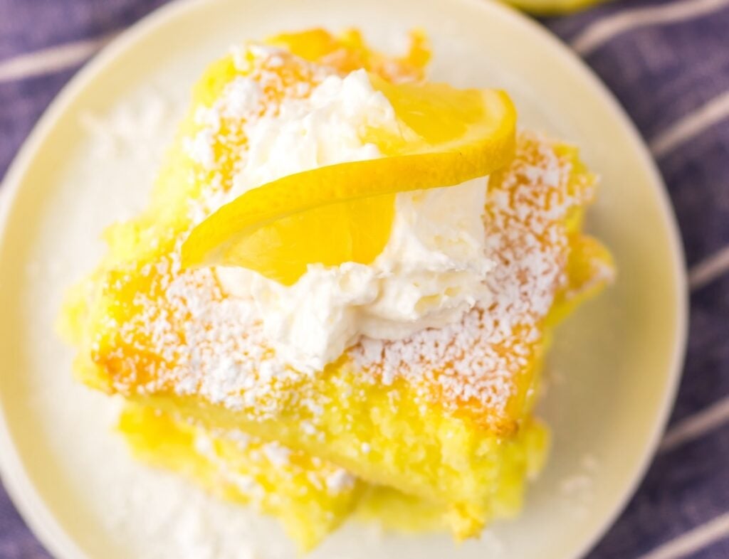 A slice of lemon cake topped with whipped cream, a lemon slice, and powdered sugar on a white plate.