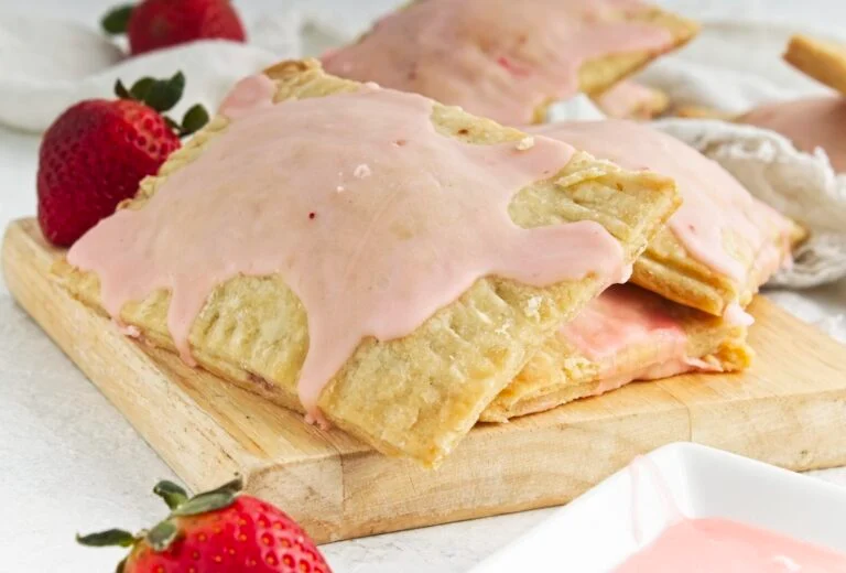 Rectangular pastries with pink icing are stacked on a wooden board with fresh strawberries beside them.