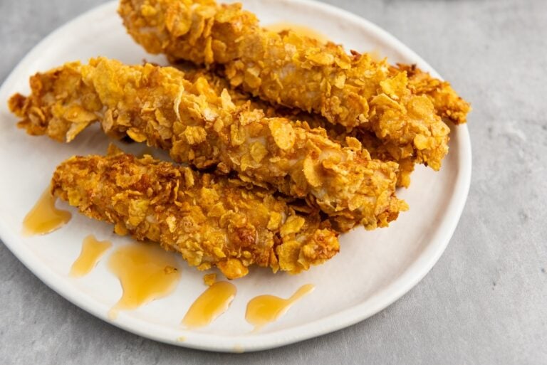Three crispy chicken tenders on a white plate, drizzled with honey.