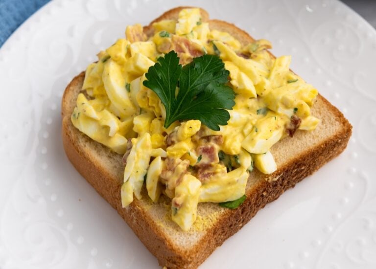 A slice of bread topped with egg salad, garnished with a sprig of parsley, on a white plate.