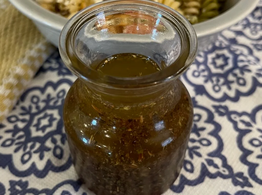 A small glass bottle filled with a brown, herbed liquid is placed on a blue and white patterned tablecloth. A portion of a bowl with mixed greens is visible in the background.