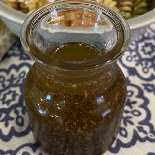A small glass bottle filled with a brown, herbed liquid is placed on a blue and white patterned tablecloth. A portion of a bowl with mixed greens is visible in the background.