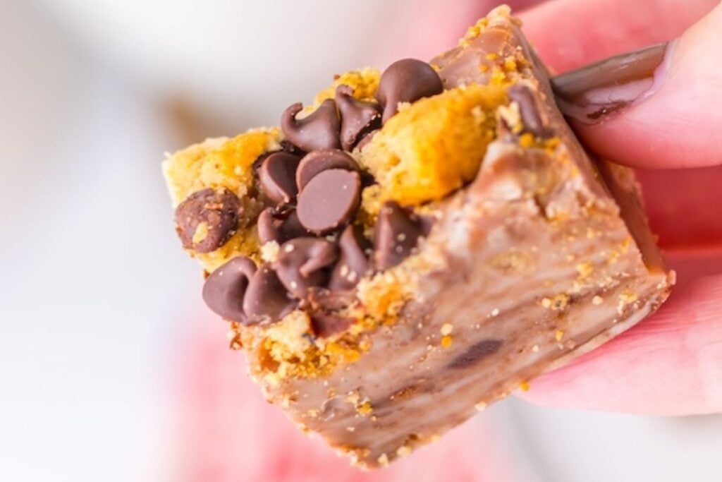 A hand holds a square dessert bar topped with chocolate chips and cookie crumbles.