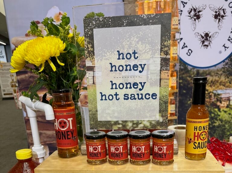Display of various "Hot Honey" and "Honey Hot Sauce" products with jars and bottles on a wooden shelf, alongside yellow flowers and a sign with the product names.