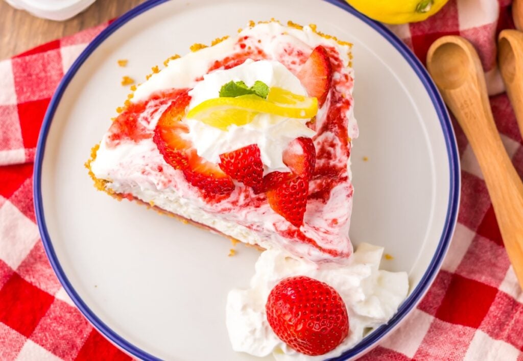 A slice of strawberry lemon pie with whipped cream, garnished with strawberry and lemon slices, on a white plate with blue rim on a red-checked tablecloth.