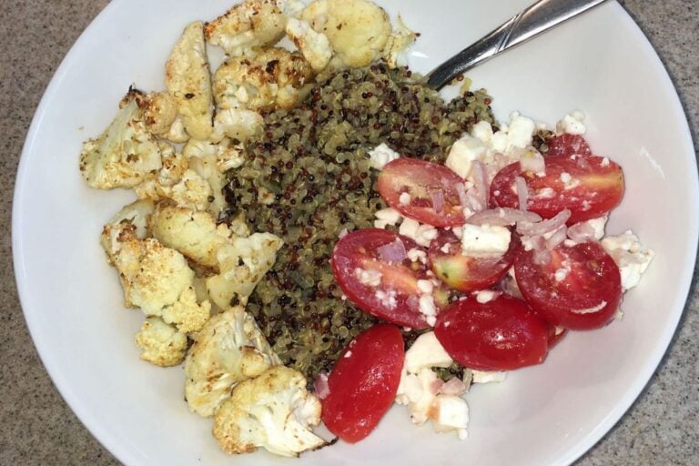 A bowl with roasted cauliflower, frozen quinoa, tomatoes, feta cheese, and onions sits invitingly with a spoon on the side.
