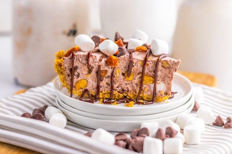 A slice of chocolate and marshmallow pie with a graham cracker crust, topped with mini marshmallows, chocolate chips, nuts, and chocolate drizzle on stacked white plates.