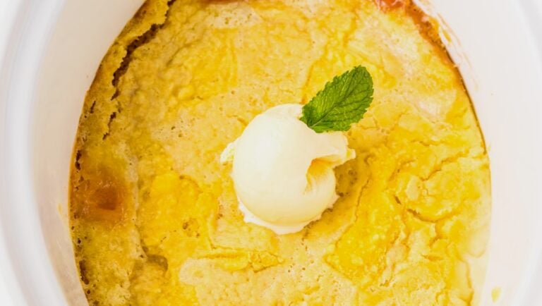 A close-up of a baked peach cobbler topped with a scoop of vanilla ice cream and a mint leaf in a white dish.