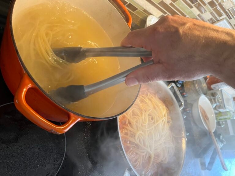 A hand uses tongs to transfer cooked spaghetti from a pot of boiling water to a pan of sauce on a stove.