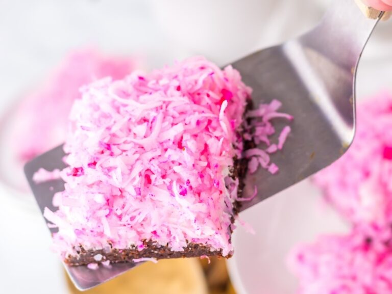 A metal spatula holds a square dessert topped with shredded pink coconut and a chocolate base.