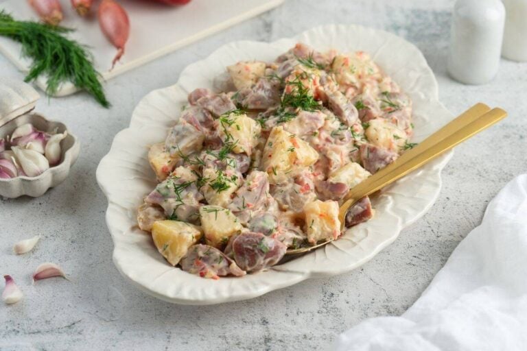 A serving dish filled with Classic Red Potato Salad with Pickles is garnished with fresh dill, with a gold-colored serving spoon resting in the dish.