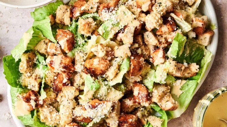 A Caesar salad topped with grilled chicken pieces, croutons, and grated cheese on a bed of romaine lettuce. Dressing is served on the side.