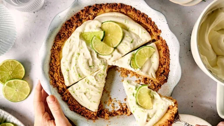 Key lime pie with a slice removed on a white plate. Topped with lime wedge and zest, the crust is golden brown. A hand touches the plate edge, and a small bowl with cream is nearby.