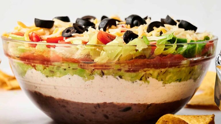 A clear glass bowl filled with layered dip, including refried beans, sour cream, guacamole, salsa, lettuce, cheese, diced tomatoes, and sliced black olives. Tortilla chips are nearby.