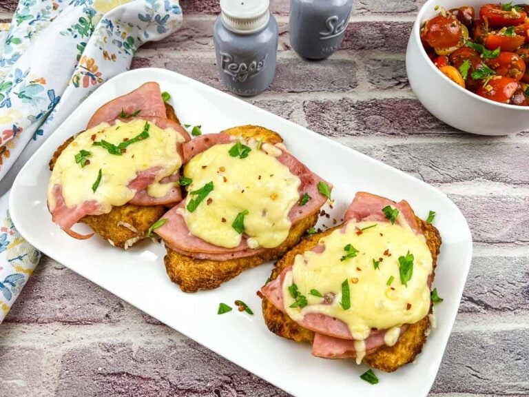 Three slices of bread topped with ham, melted cheese, and chopped herbs on a white plate, next to a bowl of tomato salad and salt and pepper shakers.