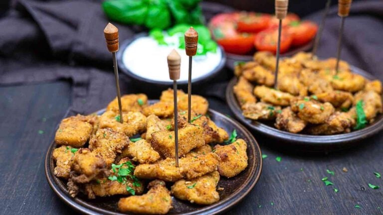 Two plates of breaded and seasoned fried chicken pieces with toothpicks, garnished with herbs, served alongside sliced vegetables and a bowl of dip.