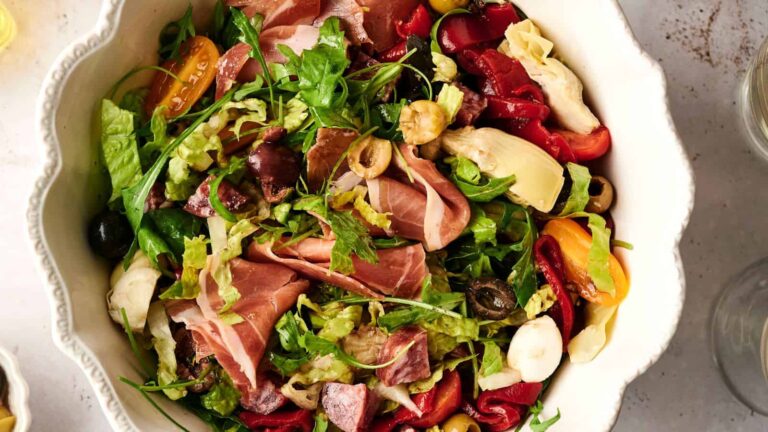 A bowl of Italian antipasto salad with prosciutto, artichoke hearts, olives, cherry tomatoes, greens, and roasted red peppers.