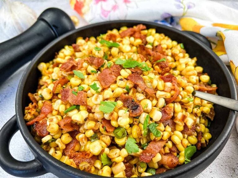 A black bowl filled with sautéed corn, crispy bacon pieces, and chopped parsley, with a spoon resting inside and a pepper grinder in the background.