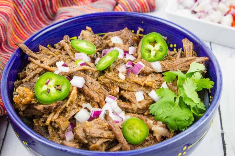 A blue bowl filled with shredded beef topped with sliced jalapeños, diced red onions, and cilantro sits on a white surface.