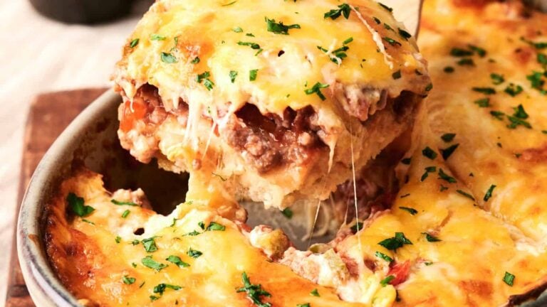 A close-up of a cheesy baked lasagna being served from a dish, with visible layers of pasta, meat, and sauce topped with melted cheese and chopped herbs.