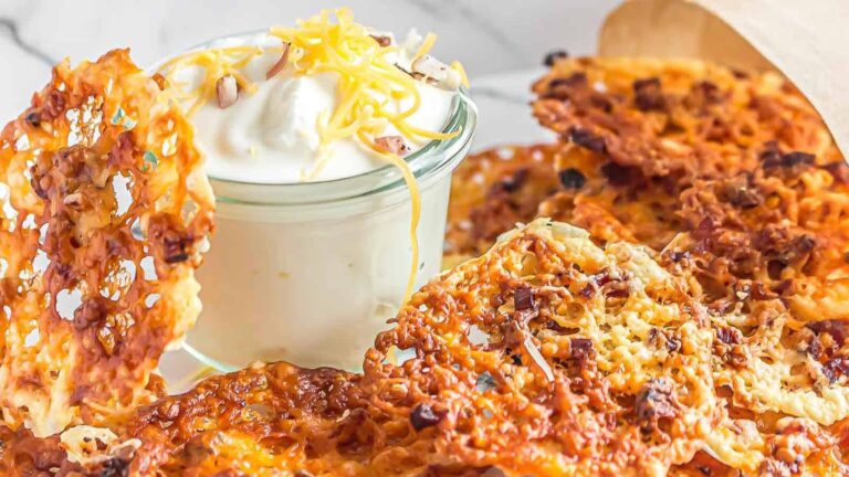A pile of crispy cheese chips with bacon sits next to a glass jar of sour cream topped with shredded cheese.