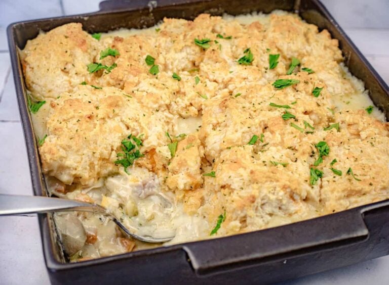 A baking dish filled with creamy chicken and biscuit casserole topped with golden biscuits and garnished with chopped parsley. A serving spoon rests inside the dish.
