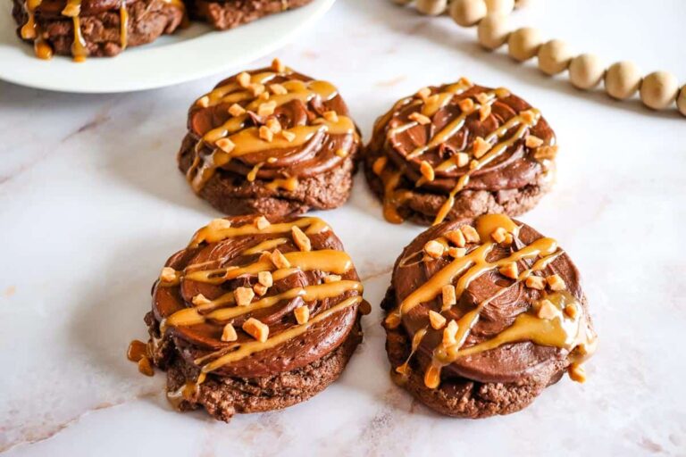 Four chocolate cookies topped with chocolate frosting, caramel drizzle, and chopped nuts sit on a white surface. A plate of additional cookies and a decorative bead garland are in the background.
