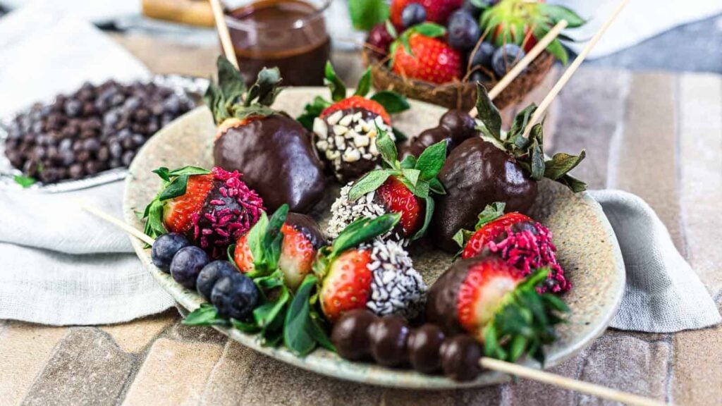 A plate with chocolate-covered strawberries, fresh blueberries, and skewered chocolate-dipped fruit, garnished with shredded coconut and colorful sprinkles.