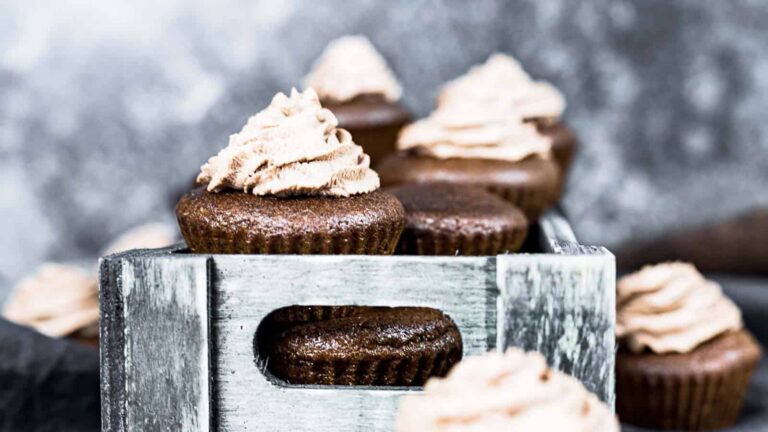 Chocolate cupcakes with swirls of light brown frosting are arranged in and around a rustic wooden box, with a blurred grey background.