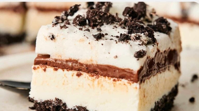 A close-up of a layered dessert with a chocolate crumb base, creamy filling, chocolate layer, whipped topping, and crushed chocolate cookies on top.