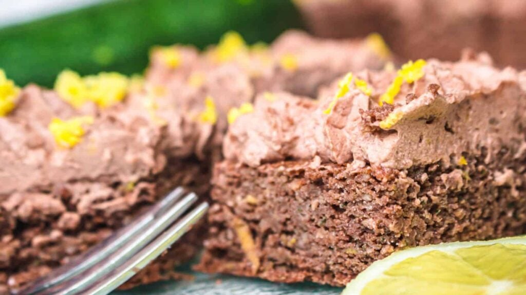 A close-up of a chocolate frosted cake slice, with a fork and a lemon wedge in the foreground and green zucchini blurred in the background.