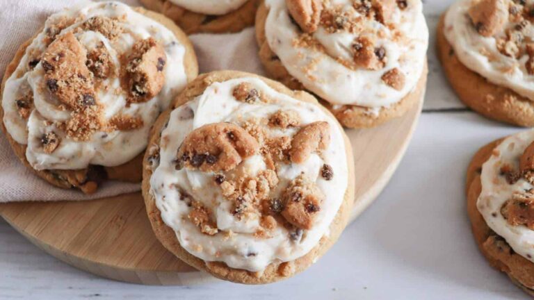 Cookies topped with creamy frosting and mini chocolate chip cookies, arranged on a wooden board and white surface.