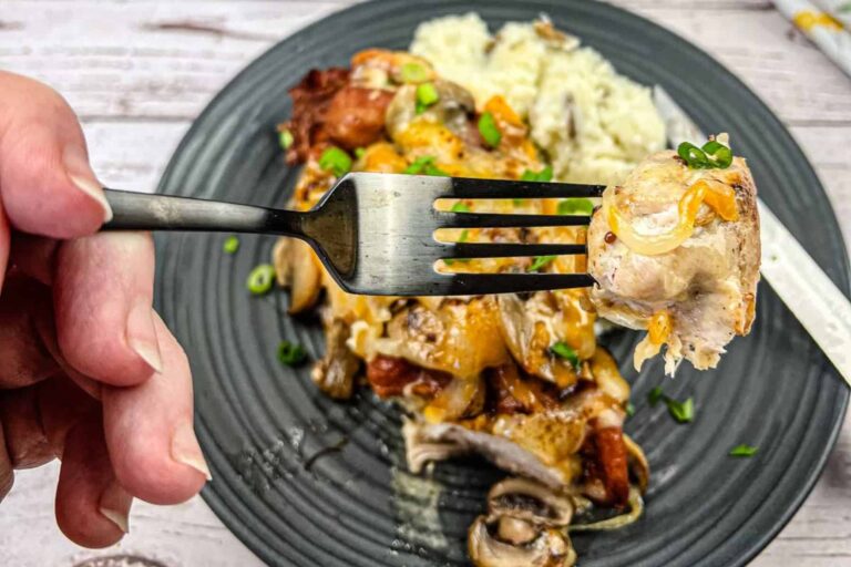 A fork holds a bite of cheesy baked chicken with mushrooms and green onions, with the rest of the meal and rice on a black plate in the background.