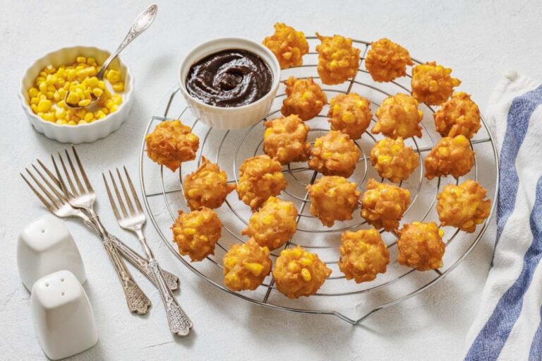 A cooling rack with golden corn fritters, a bowl of barbecue sauce, a bowl of corn kernels, three forks, and salt and pepper shakers on a white surface.