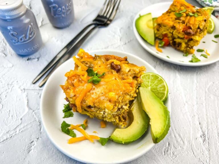 A plate with a slice of cheesy casserole, avocado slices, lime wedges, and garnish; a similar plate and utensils are in the background near salt and pepper shakers.