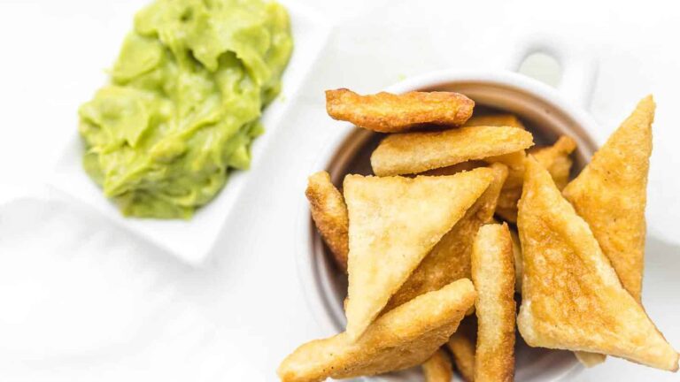 A white bowl filled with fried triangular pieces of bread is next to a small rectangular dish of guacamole on a white surface.