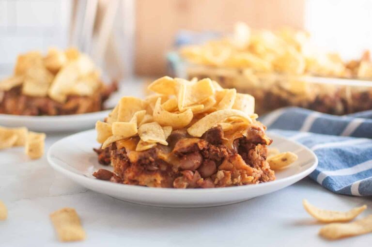A slice of Frito pie topped with corn chips on a white plate, with more Frito pie and a blue-striped napkin in the background.