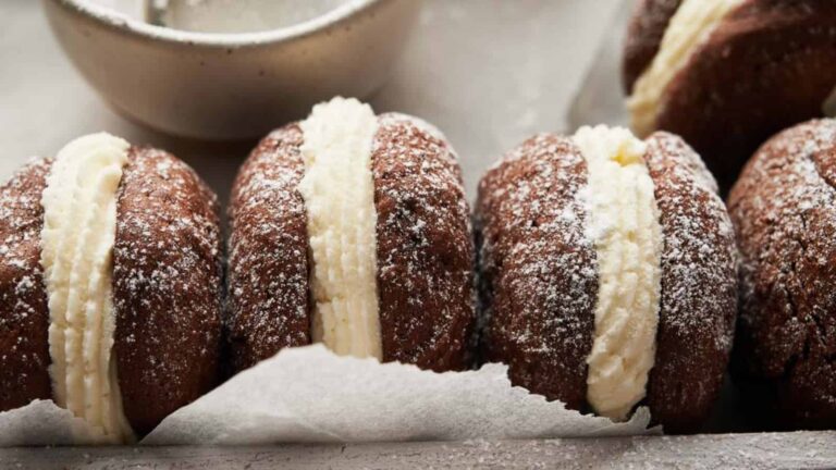 Four chocolate sandwich cookies filled with cream and dusted with powdered sugar are lined up on parchment paper, with part of a bowl in the background.